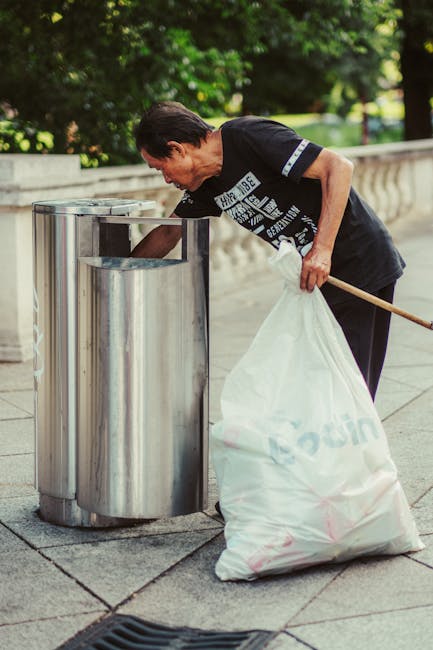 A middle-aged man with dark hair, dressed in a black T-shirt with white text and dark trousers, is actively disposing of waste at a public outdoor recycling station. He is bent slightly forward, holdi