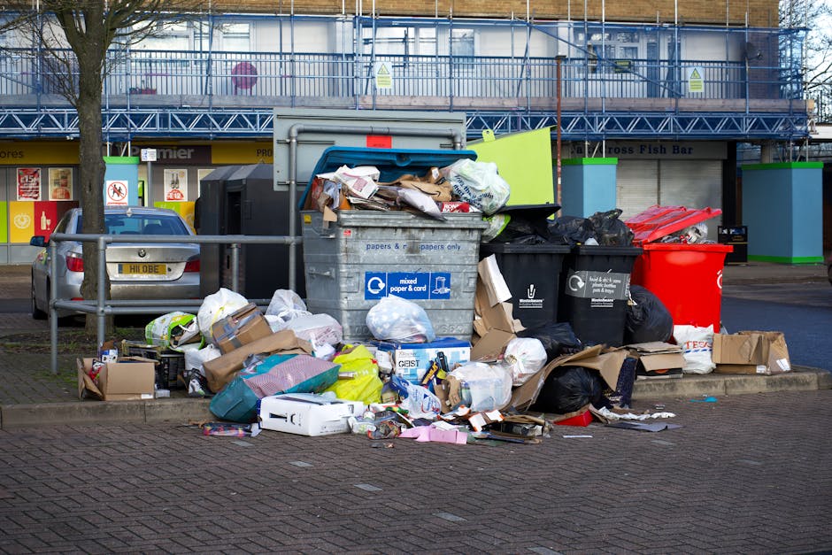 A roadside pile of waste and rubbish, including black and grey garbage bags, a yellow plastic container, and a discarded dirty beige sofa cushion with visible fabric and stuffing, all resting on a pav